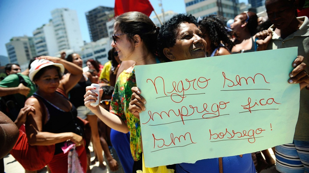 Mulher negra segura um cartaz feito à mão em uma manifestação: "Negro sem emprego fica sem sossego". Ela sorri. Há outras pessoas na foto, uma bandeira ao fundo.