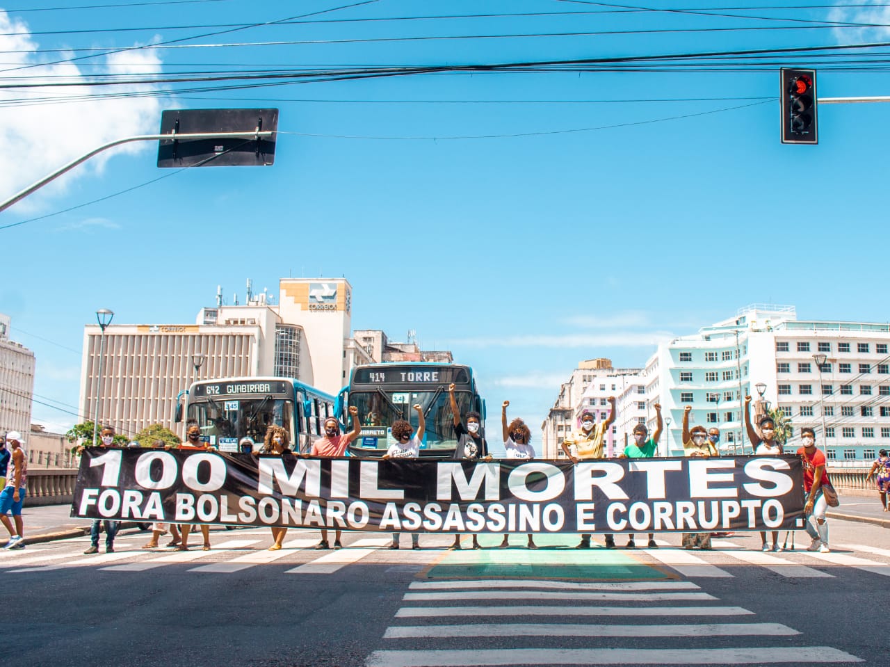 Uma faixa preta com letras brancas é aberta em uma rua, diante do trânsito. Escrito 100 mil mortes. Bolsonaro assassino.
