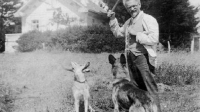 Trotsky está de pé, com um cachorro preso por uma corda. ele olha em direção a câmera. e segura uma pedaço de madeira. Ao fundo, uma casa.