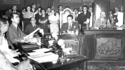 Um salão de reuniões. Uma cadeira ao centro, com quem preside a sessão. à esquerda, um homem discursa. Ao fundo, várias pessoas assistem à sessão.
