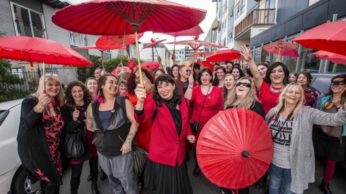 cerca de 30 mulheres posam para foto em uma rua. Elas estão sorridentes e algumas estão com sombrinhas de papel, contra o sol. A maioria usa camisas na cor vermelha.
