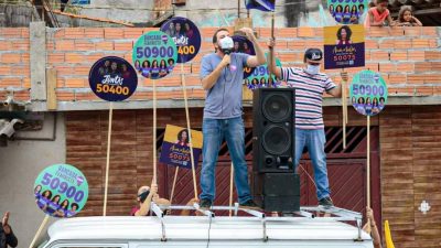 Guilherme Boulos discursa em cima de uma kombi, em atividade de campanha. Ele usa máscaras. Em torno dele, cinco ou seis pirulitos de campanhas para vereador(a).