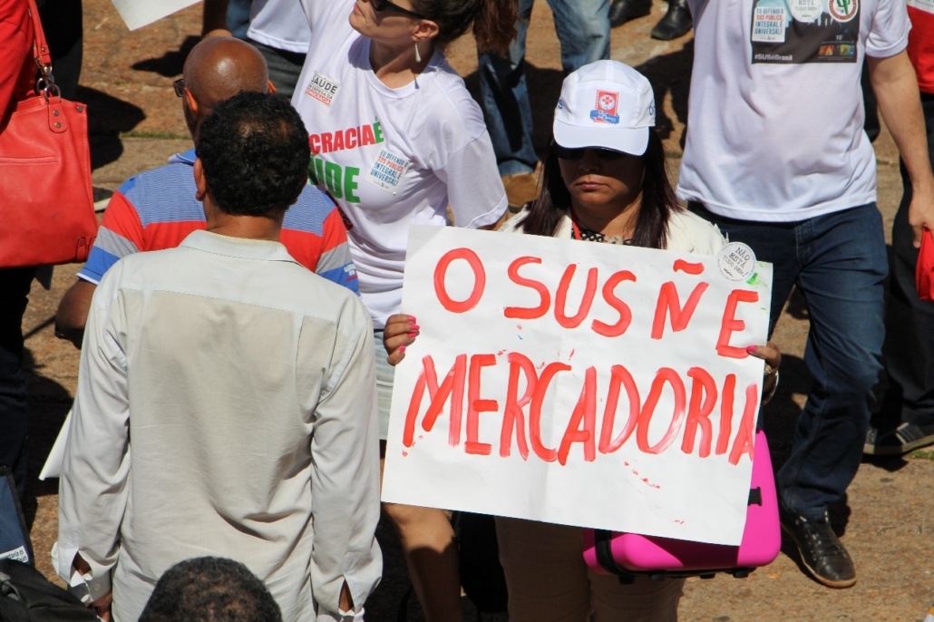 Imagem de um protesto. Uma servidora da saúde, com um boné, segura um carta: O SUS Ñ É MERCADORIA