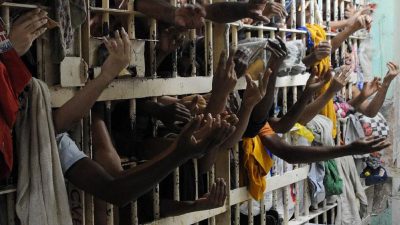 Foto de uma grade de uma cela, com superlotação. Vários detentos colocam as mãos para fora.