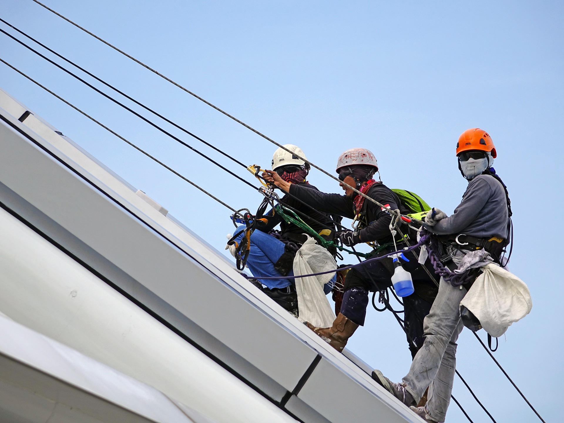 três trabalhadores estão escalando uma construção, segurando em cabos de aço. Eles usam capacetes e botas. Um deles olha em direção à câmera.