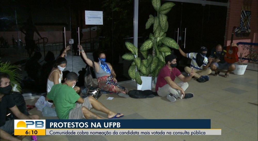 Reprodução da TV. Duas jovens estão acorrentadas aos puxadores da porta de vidro da reitoria. Outros estudantes também estão juntos, sentados no chão.