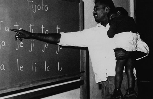 Um homem segura no colo uma criança e aponta para sílabas em um quadro escolar