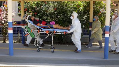 Dois profissionais da saúde carregam um paciente em uma maca. Eles usam mascaras e roupas de proteção brancas. O paciente é um homem, usa bermuda e camisa do flamengo.