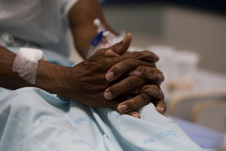 Foto mostra mãos cruzadas de uma mulher idosa negra em um hospital