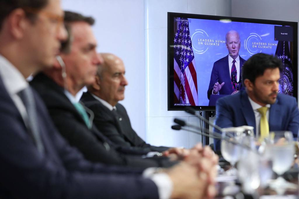 Bolsonaro e ministros estão sentados, com fones de ouvido. Ao fundo, a bandeira dos EUA e Joe Biden, discursando na Cúpula do Clima