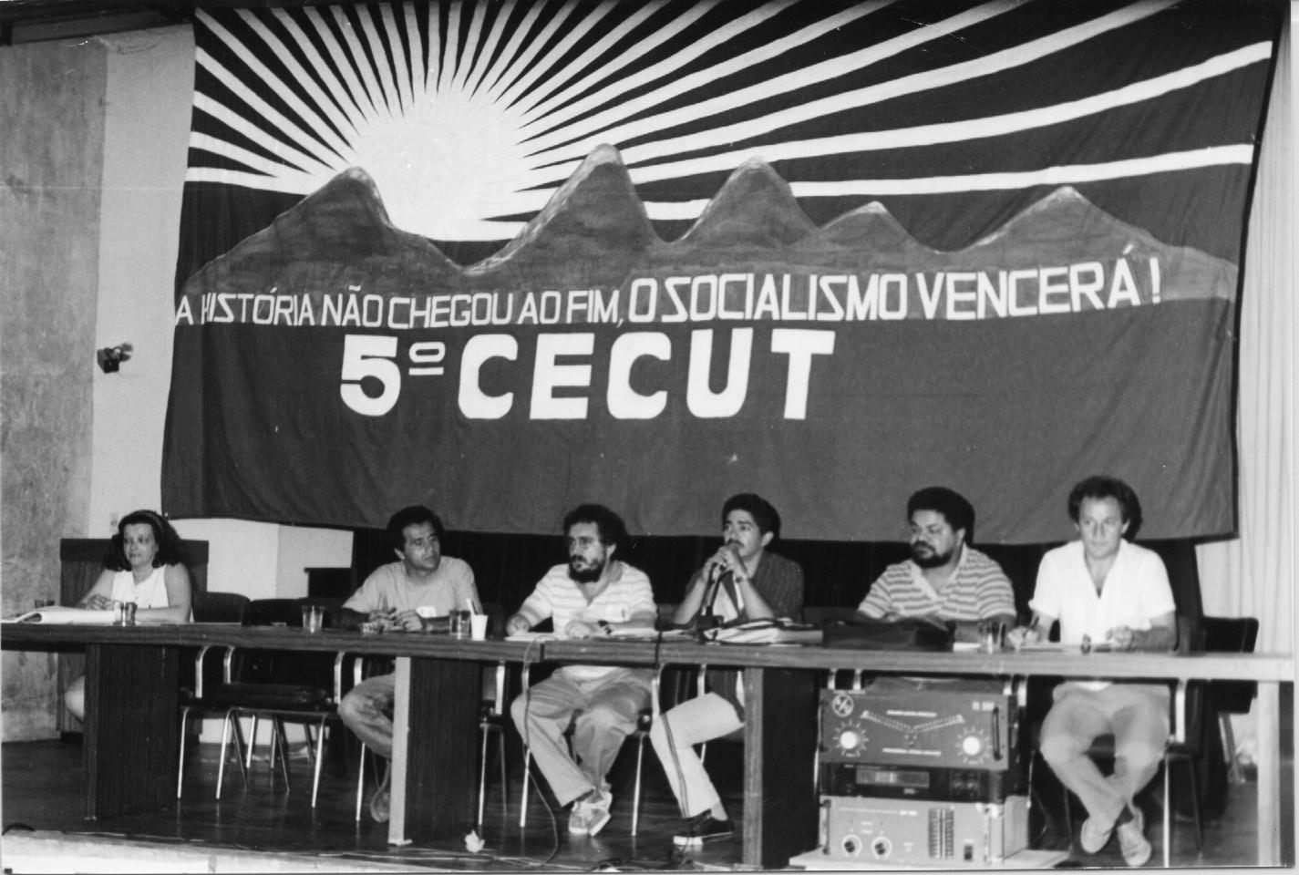 Lays na mesa do 5 Congresso Estadual da CUT. Ao seu lado, cinco homens. Na faixa, acima, está escrito. A história não chegou ao fim. O socialismo vencerá! 
