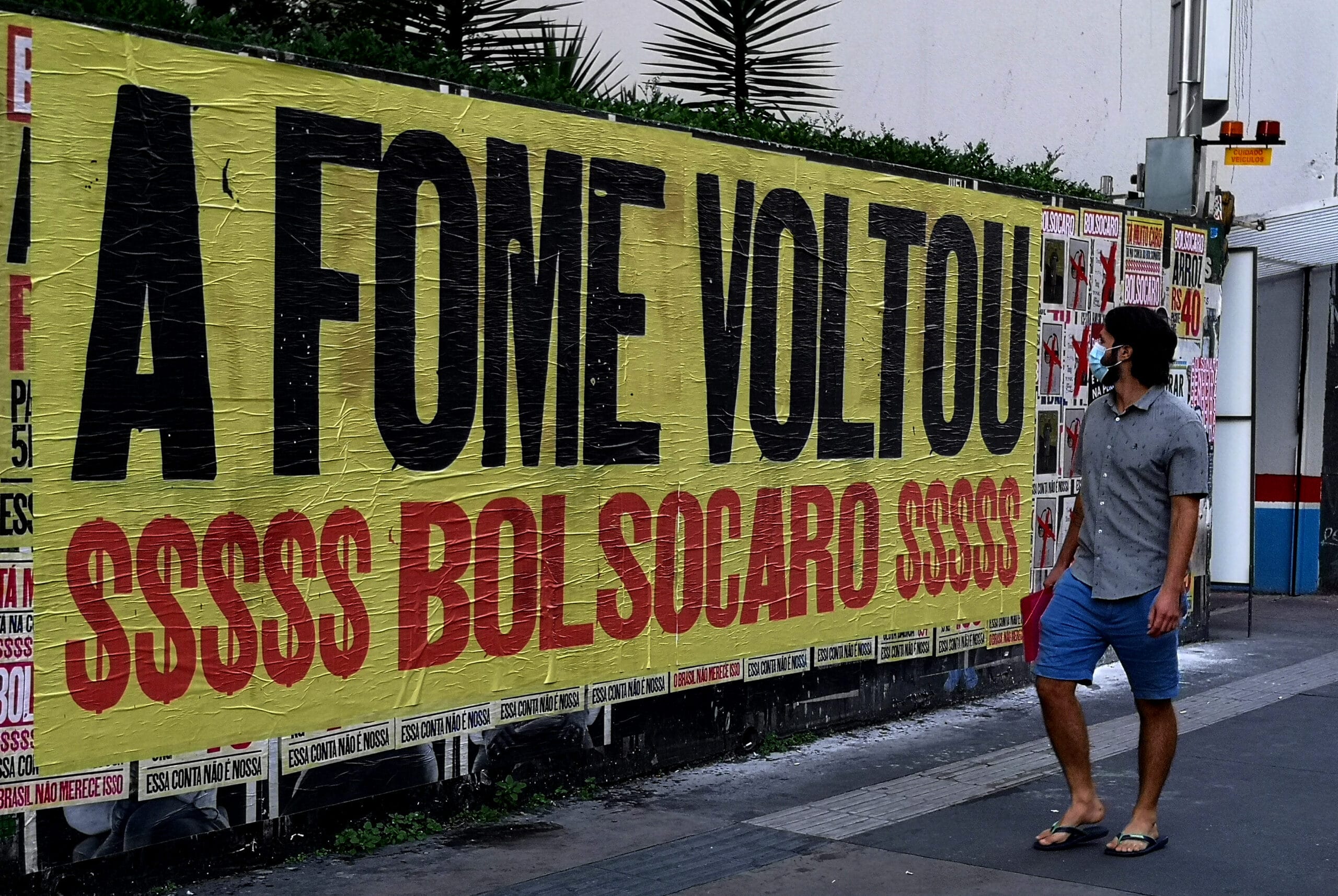 Cartaz colado na rua, com a frase: "A FOME VOLTOU!" abaixo: $$$$$ BOLSOCARO $$$$$. Um homem passa e observa o mural.