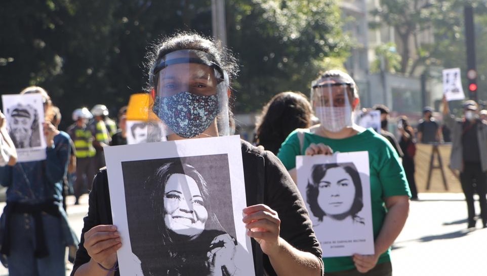 Manifestantes exibem cartazes com fotos de vítimas da covid. São mulheres, usam face shield, máscara e estão a uma distância segura