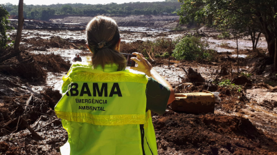 MUlher com colete amarelo, escrito IBAMA Emergência Ambiental nas costas. Ela fotografa o mar de lama em Brumadinho