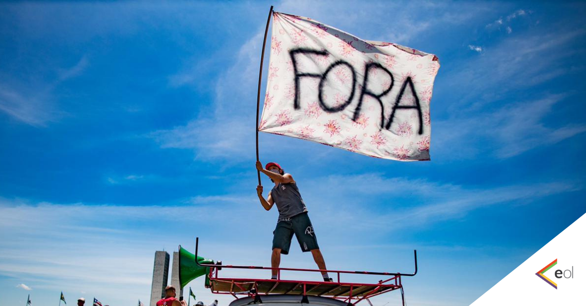 Homem segura ao alto uma bandeira branca onde se lê a palavra FORA.Ele está em cima de um carro. ao fundo, o céu azul de Brasília.