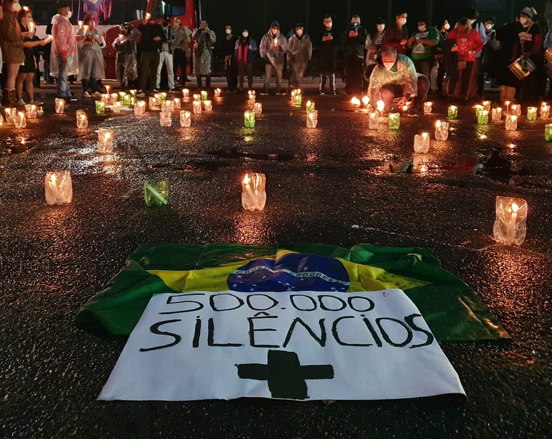 Foto mostra velas acesas e um cartaz no chão, escrito 500.000 silêncios. com uma cruz e uma bandeira do Brasil