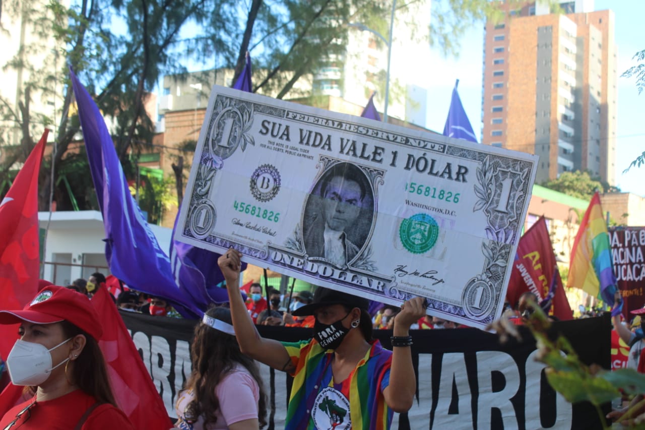Manifestante carrega reprodução de nota de um dólar com a foto de Bolsonaro, e na nota, a frase? Sua vida vale 1 dólar?. Ela usa uma bandeira LGBT pendurada, como um lenço e a camisa do MST. A seu redor, faixas e bandeiras.