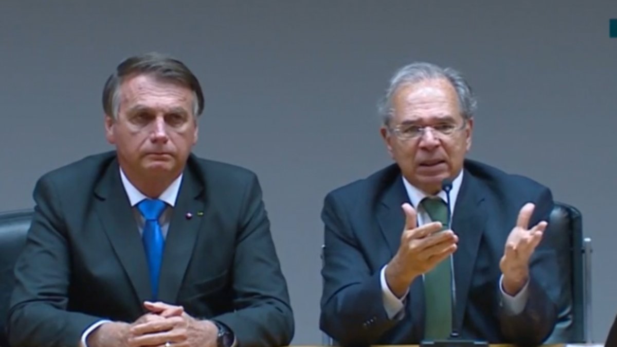 Bolsonaro está à esquerda, de terno escuro e gravata azul. Guedes, ao seu lado, está falando, com gravata cinza e com as mãos para a frente.