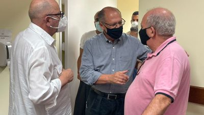 Alckmin cumprimenta dois sindicalistas. Ele usa máscara e calças pretas e camisa social de mangas compridas azul. Os dois sindicalistas estão de camisa branca social, à esquerda, e camisa polo, rosa, à direita. Os três são brancos e calvos..