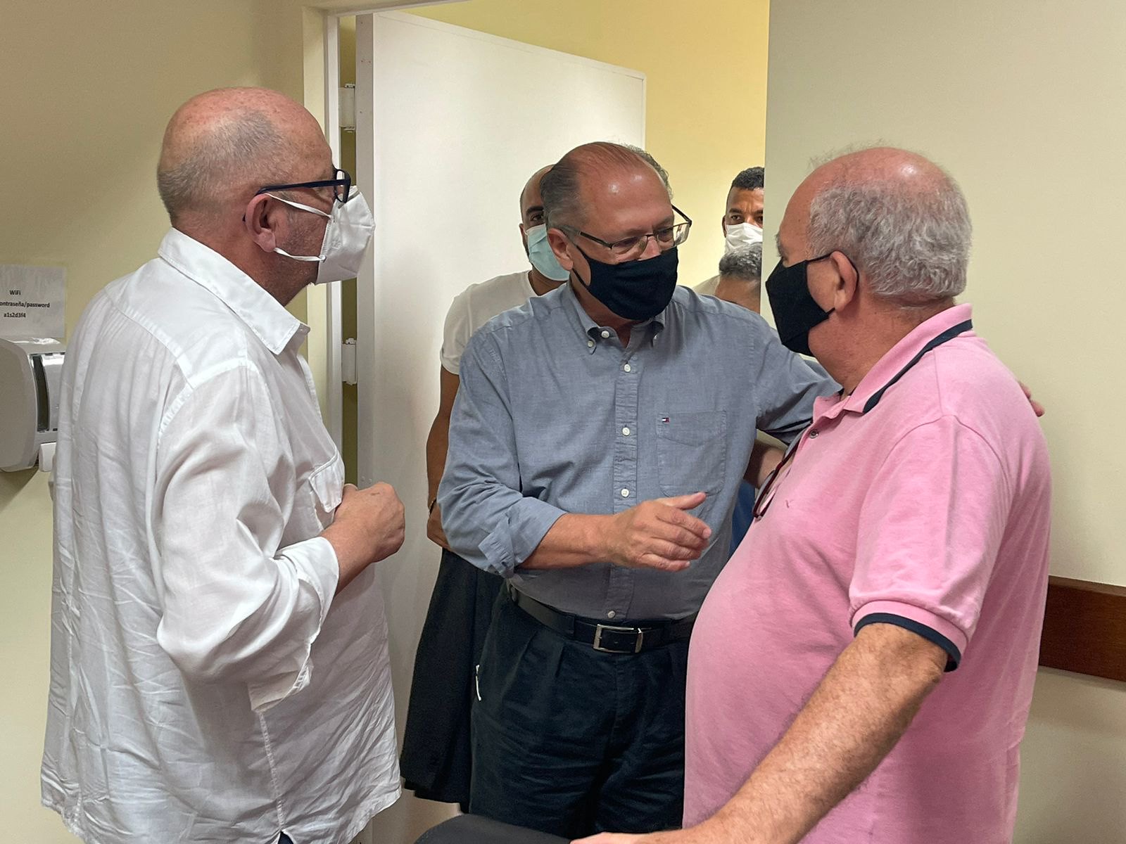 Alckmin cumprimenta dois sindicalistas. Ele usa máscara e calças pretas e camisa social de mangas compridas azul. Os dois sindicalistas estão de camisa branca social, à esquerda, e camisa polo, rosa, à direita. Os três são brancos e calvos..