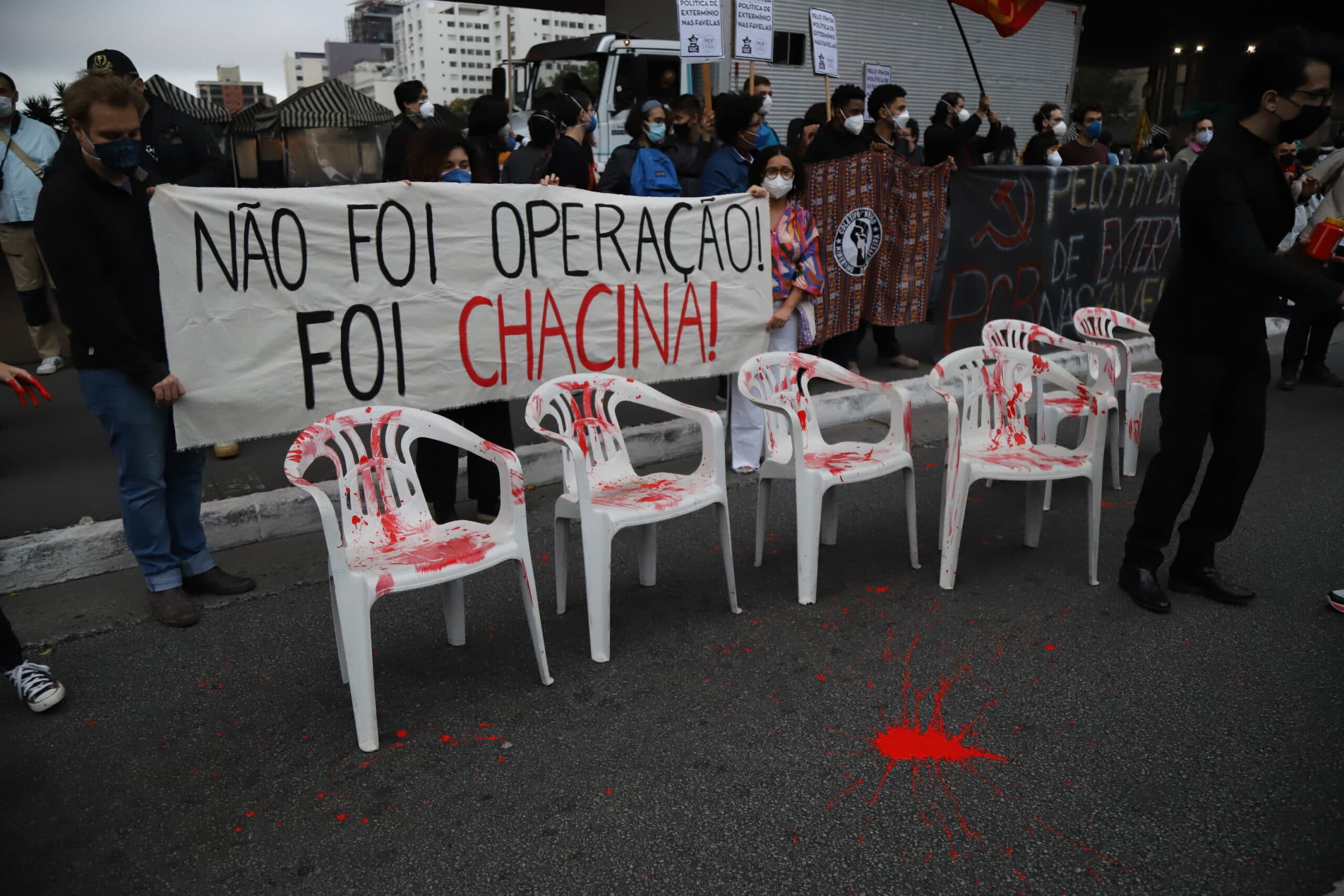 foto do ato mostra cadeiras de plástico manchadas de tinta vermelha. atrás, uma faixa; não foi operação, foi chacina.