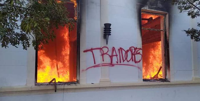 Foto mostra a fachada de um prédio público, com as chamas em seu interior, que são vistas pelas duas janelas. Na fachada, a pichação em vermelho: traidores.