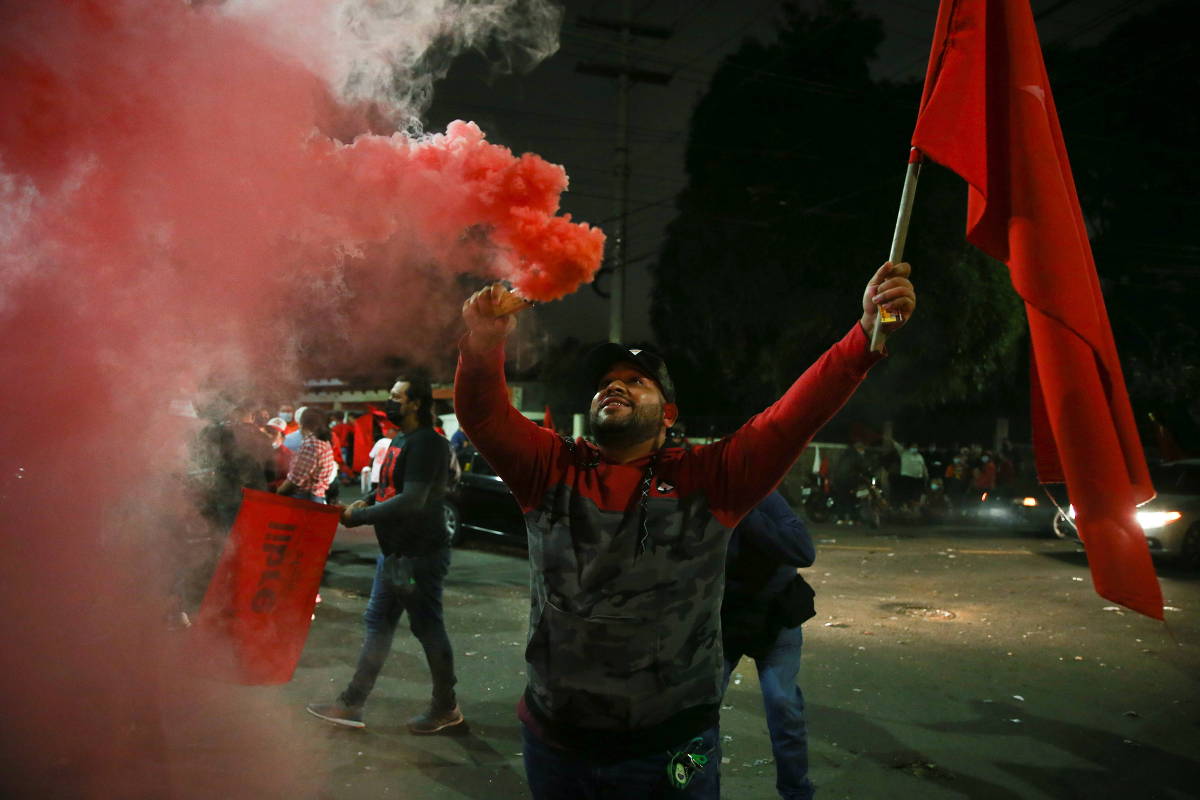 Um apoiador comemora, com uma bandeira vermelha na mão esquerda e com a outra, soltando uma fumaça vermelha.