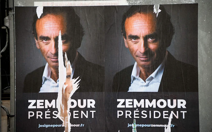 Cartazes de rua com a foto do candidato e abaixo a frase Zemmour Président. Ele usa terno casaco preto sobre uma camisa azul clara. é um homem branco, em torno de 60 anos, calvo.