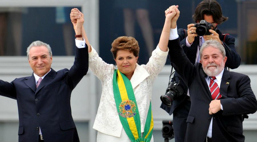 Temer, Dilma (com a faixa presidencial) e Lula, na cerimônia de transmissão do cargo, em 2015. Os três estão na rampa do palácio e com as mãoes erguidas.