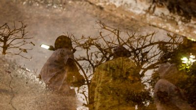 Foto mostra no reflexo de uma poça dágua, três manifestantes caminhando e acima deles, galhos de uma árvore, sem folhas.