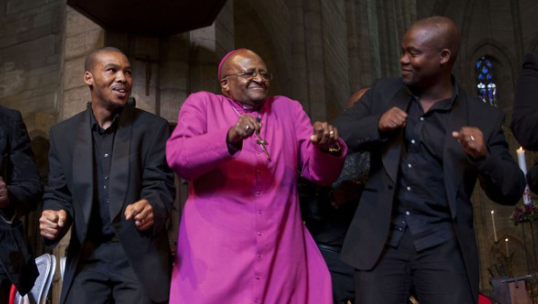Desmond Tutu está dançando, no altar de uma igreja. Ao seu lado, dois homens de terno. Ele usa uma túnica roxa. Todos são negros.