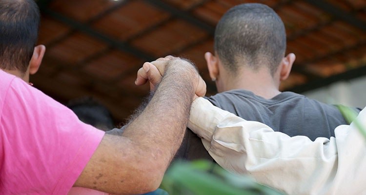 foto mostra detalhe pessoas de costas, de mãos dadas, como se estivessem orando. Um deles tem a cabeça raspada