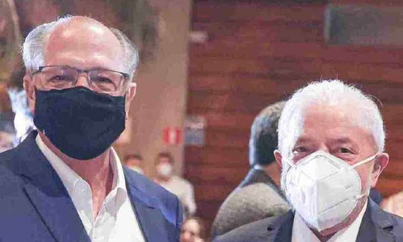 Lula e Alckmin, lado a lado, em jantar. Alckmin é homem branco, usa óculos, máscara preta, é calvo. Lula é homem branco, cabelos grisalhos, e máscara branca. Alckmin é mais alto que Lula. Os dois usam terno. Alckmin não usa gravata.