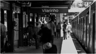 Estação Vilarinha do metrô de BH. Foto mostra passageiros de costas junto a composição e em destaque a placa com o nome da estação. A foto é em preto e branco