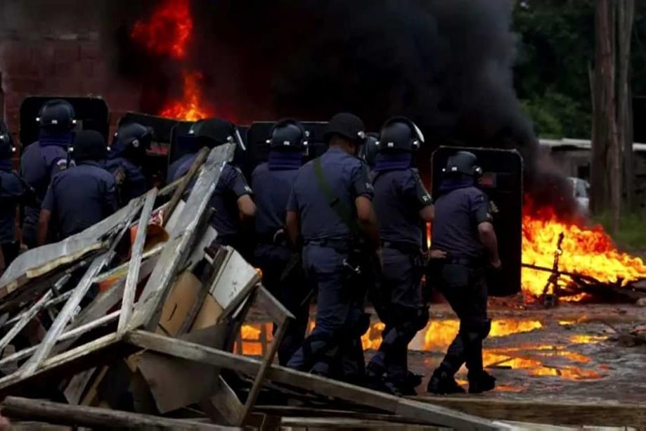 Policiais avançam em formação. A frente, chamas e fumaça. Atrás deles, pedaços de madeira de uma das casas, destruída.
