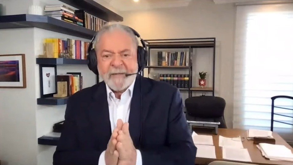 Foto mostra Lula na entrevista. Ele usa um fone de ouvido e microfone. Veste um terno azul marinho e camisa social branca. Está em um escritóirio, com algumas prateleiras e estantes. uma mesa ao fundo e uma cadeira preta.
