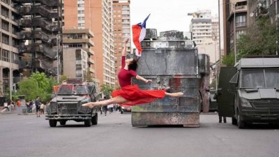 foto mostra uma bailarina, com roupa vermelha, saltando. Ao fundo, militares em um tanque, observam a dança.