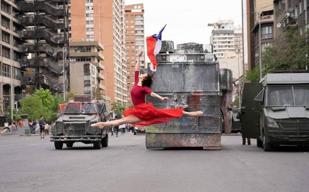 foto mostra uma bailarina, com roupa vermelha, saltando. Ao fundo, militares em um tanque, observam a dança.