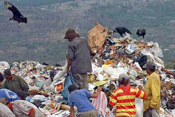 Foto mostra pessoas em um lixão. São 3 homens negros, de costas. Ao fundo, acima da montanha de lixo, urubus.
