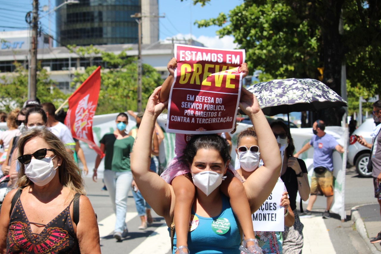 Mulher branca de máscara com criança nos ombros segura um cartaz com "Estamos em greve: em defesa do serviço público, por direitos e contra a terceirização" em meio à multidão