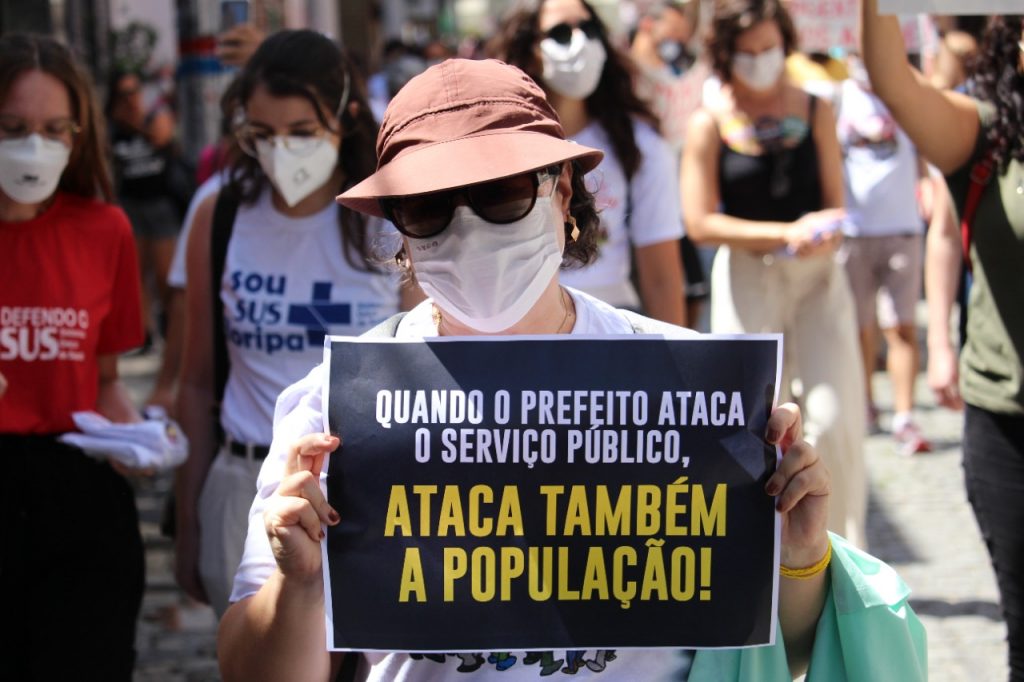 Mulher de máscara, chapéu e óculos segura cartaz "Quando o prefeito ataca o serviço público, ataca também a população!"