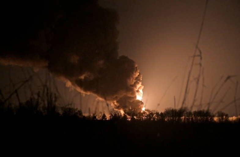 Foto noturna, mostra uma nuvem de fumaça ao longe iluminada por uma explosão do lado direito.