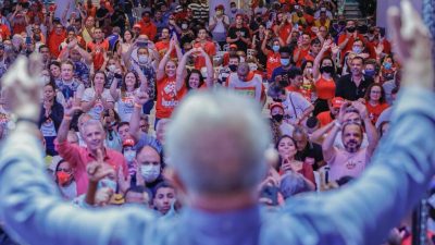 Lula, de costas, acena para a o público a sua frente. Centenas de pessoas fazem sinal de coração com as mãos