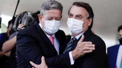 Lira e Bolsonaro se abraçam. Eles estão rindo. Usam máscaras