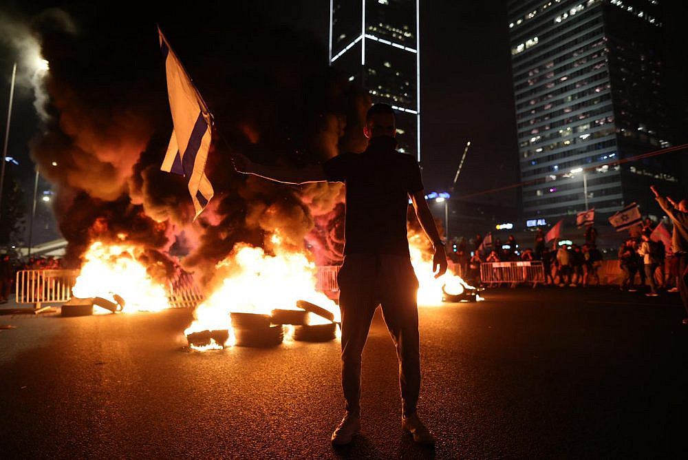 Um manifestante fica em frente a pneus em chamas na rodovia Ayalon, Tel Aviv. Dezenhas de milhares de manifestantes foram às ruas após o primeiro ministro Benjamin Netanyahu demitir o ministro da Defesa, Yoav Gallant, depois que ele pediu uma pausa na reforma judicial pelo governo